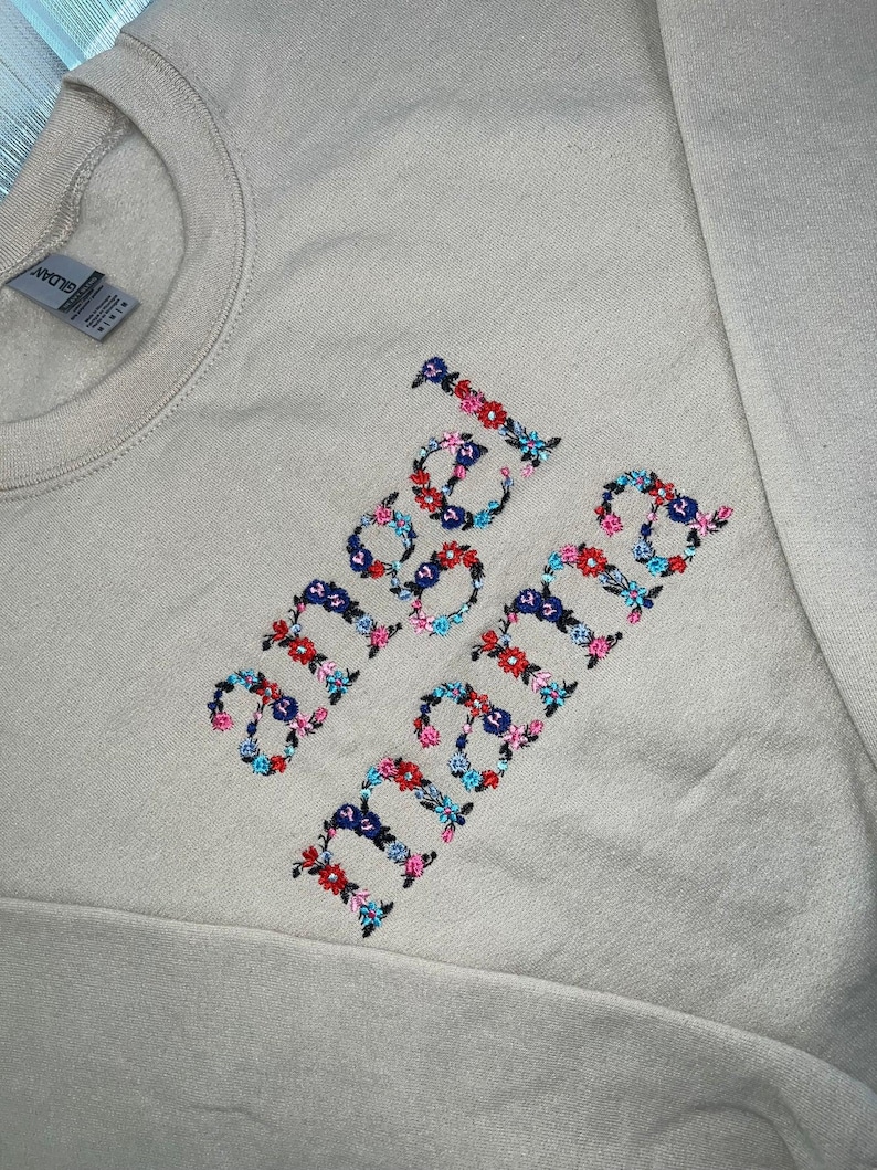 May include: A beige sweatshirt with the embroidered words "angel mama" in colorful floral lettering. The embroidery features a mix of red, blue, and pink flowers, creating a vibrant contrast against the neutral background. The sweatshirt has a crew neck and long sleeves.