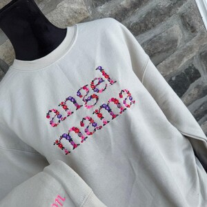 May include: Cream-colored sweatshirt with the embroidered text "angel mama" in a floral design. The sleeve cuff features the name "Sutton" in pink embroidery. The sweatshirt is displayed on a black mannequin head.