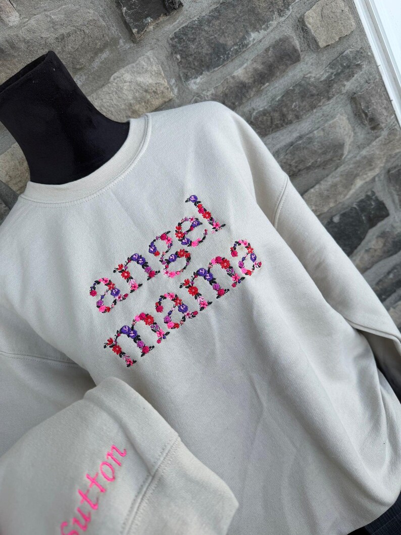 May include: Cream-colored sweatshirt with the embroidered text "angel mama" in a floral design. The sleeve cuff features the name "Sutton" in pink embroidery. The sweatshirt is displayed on a black mannequin head.