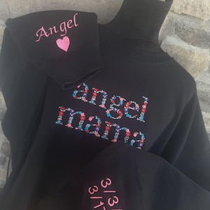 May include: Black turtleneck sweatshirt with embroidered text and floral design. The word "Angel" is embroidered on the sleeve with a pink heart. The words "Angel mama" are embroidered on the front. Dates are embroidered on the other sleeve.