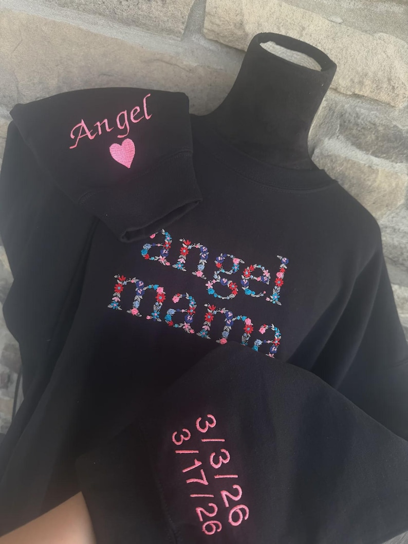 May include: Black turtleneck sweatshirt with embroidered text and floral design. The word "Angel" is embroidered on the sleeve with a pink heart. The words "Angel mama" are embroidered on the front. Dates are embroidered on the other sleeve.