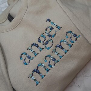 May include: A beige sweatshirt with the words "angel mama" embroidered in blue and white floral letters. The sweatshirt has a crew neck and long sleeves.