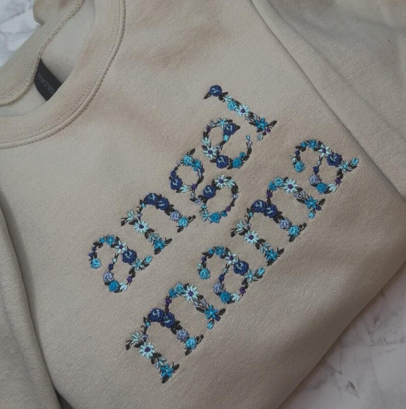 May include: A beige sweatshirt with the words "angel mama" embroidered in blue and white floral letters. The sweatshirt has a crew neck and long sleeves.
