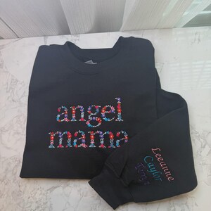 May include: Black sweatshirt with the words "angel mama" embroidered in a floral pattern. The right sleeve has the names "Leeanne Caylor Terryn" embroidered in pink, teal, and purple.