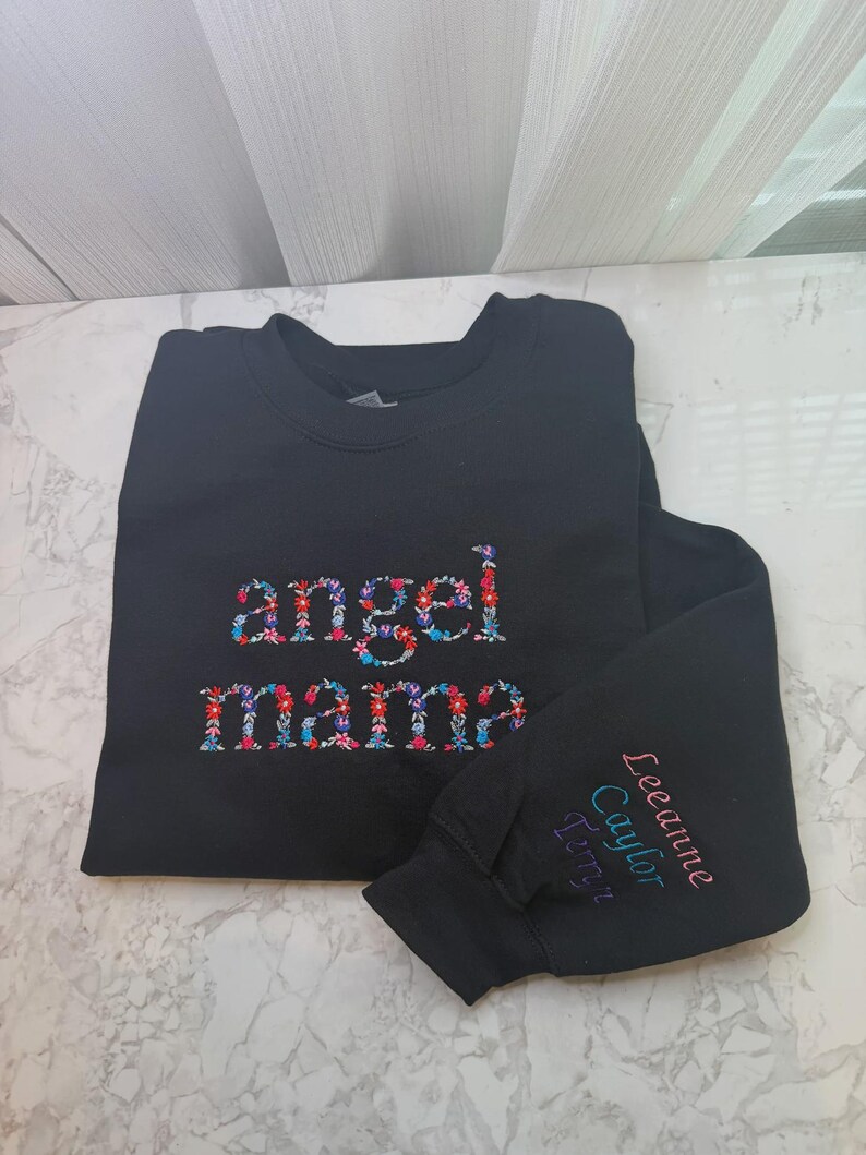 May include: Black sweatshirt with the words "angel mama" embroidered in a floral pattern. The right sleeve has the names "Leeanne Caylor Terryn" embroidered in pink, teal, and purple.