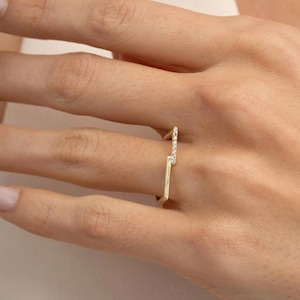 Puede incluir: Un anillo de oro con un diseño geométrico abierto que presenta dos barras paralelas conectadas en ángulo. Una barra está engastada con una hilera de pequeñas piedras claras y brillantes, mientras que la otra es de oro pulido y liso.