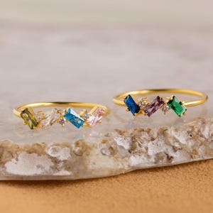 Peut inclure: Deux bagues dorées, chacune ornée d'une rangée de pierres précieuses de taille baguette colorées. L'une a des pierres vertes, claires, bleues et roses. L'autre a des pierres bleues, violettes et vertes. De petites pierres claires sont également présentes.
