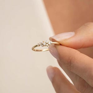 Puede incluir: Un anillo fino de oro con un grupo de cinco pequeñas piedras claras de talla redonda, dispuestas en una formación delicada y asimétrica en la parte superior de la banda. El metal tiene un acabado pulido y reflectante.