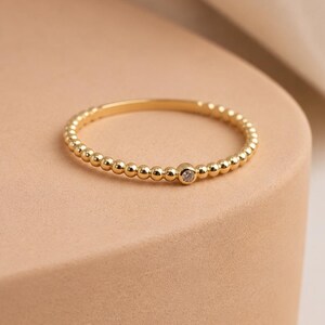Puede incluir: Un anillo fino de oro que presenta una banda continua de pequeñas cuentas metálicas uniformes. Una única piedra preciosa redonda y transparente está engastada en el centro de la banda de cuentas, creando un aspecto delicado y refinado.