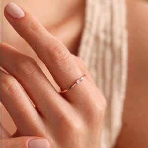 Peut inclure: Une fine bague en or rose dotée d'une unique petite pierre précieuse rectangulaire de couleur rose clair, sertie horizontalement. L'anneau en métal est lisse et fin, présentant un design épuré qui met en valeur la pierre délicate.