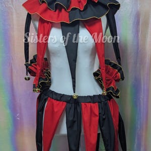 May include: A jester-style costume set featuring a red and black color-blocked design with gold trim. The set includes a ruffled collar, long pointed sleeves, and a skirt with pointed panels, all accented with small gold bells. Text reads Sisters of the Moon.