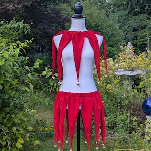 May include: A red jester costume displayed on a mannequin. The costume includes a collar, skirt, and dangling pointed pieces. Small gold bells are attached to the skirt and collar. The text "Sisters of the Moon" is at the bottom.