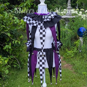 May include: A jester-style costume featuring a black and white checkered pattern combined with solid purple panels. The garment includes a ruffled collar, long sleeves with ruffled cuffs, and a pointed hemline adorned with small metal bells.