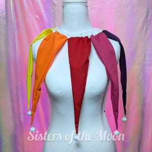 May include: A multi-colored jester collar featuring five long, pointed fabric panels in yellow, orange, red, pink, and dark purple. Each panel is tipped with a small white bell. The collar is displayed on a white mannequin with the text Sisters of the Moon.