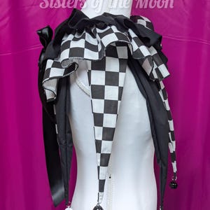May include: A black and white checkered jester collar with a ruffled design. The collar has long, pointed ends with small bells and is displayed on a white mannequin. The text "Sisters of the Moon" is visible.