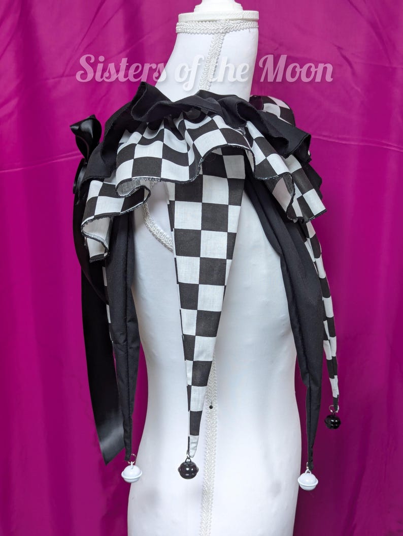 May include: A black and white checkered jester collar with a ruffled design. The collar has long, pointed ends with small bells and is displayed on a white mannequin. The text "Sisters of the Moon" is visible.