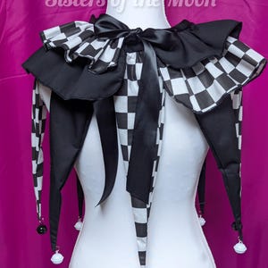 May include: A black and white chequered jester collar with a black satin bow and long, pointed tails. The collar has a ruffled edge and small bells at the tips of the tails. The text "Sisters of the Moon" is visible.