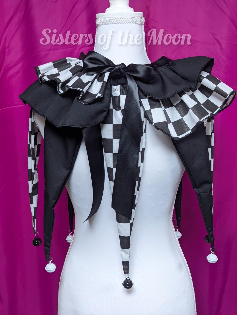 May include: A black and white chequered jester collar with a black satin bow and long, pointed tails. The collar has a ruffled edge and small bells at the tips of the tails. The text "Sisters of the Moon" is visible.
