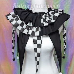 May include: A black and white chequered jester collar with a ruffled design. The collar has long, pointed ends with small bells attached. The text "Sisters of the moon" is visible in the background. This costume accessory is displayed on a white mannequin.