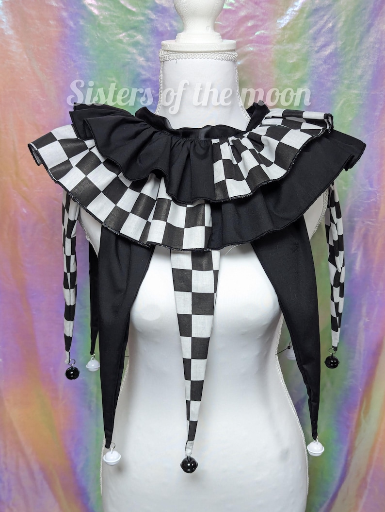 May include: A black and white chequered jester collar with a ruffled design. The collar has long, pointed ends with small bells attached. The text "Sisters of the moon" is visible in the background. This costume accessory is displayed on a white mannequin.