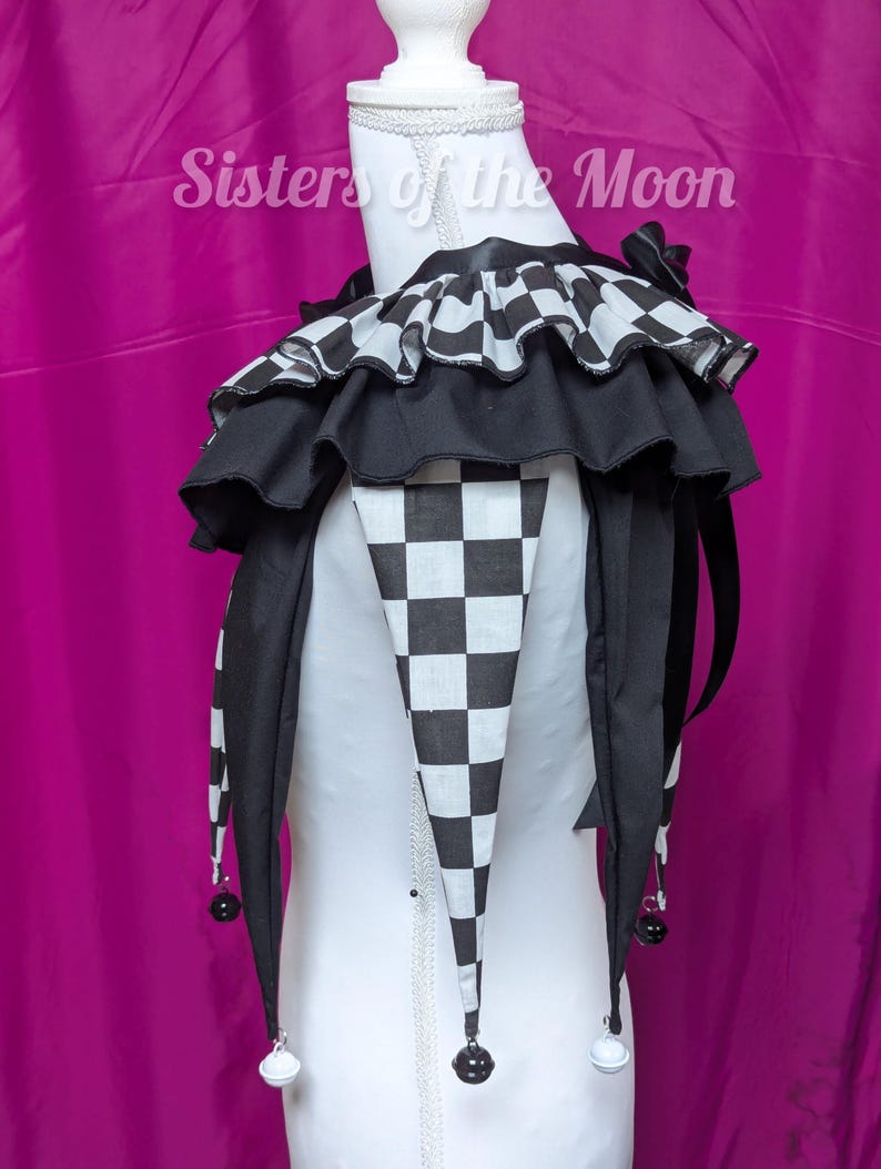 May include: A black and white jester collar with a checkered design and ruffled trim. The collar features long, pointed ends with small bells. The text "Sisters of the Moon" is visible in the background.