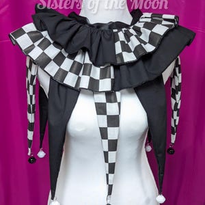 May include: A black and white chequered jester collar with a frilled edge and long, pointed ends. The collar is displayed on a white mannequin and has small bells at the tips of the points. The background is a solid magenta colour.