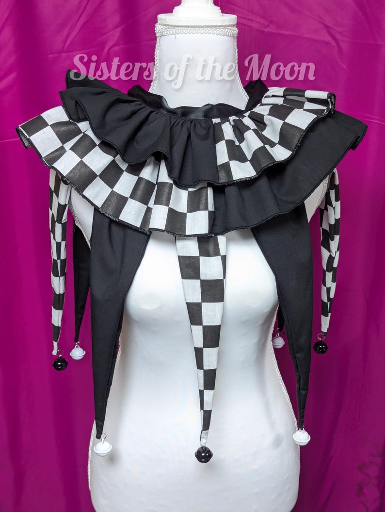 May include: A black and white chequered jester collar with a frilled edge and long, pointed ends. The collar is displayed on a white mannequin and has small bells at the tips of the points. The background is a solid magenta colour.