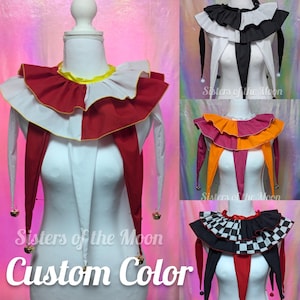 May include: A multi-layered fabric neck ruff featuring a ruffled collar with long pointed drapes and small metallic bells at the tips. The accessory is displayed in various color combinations including red and white, black and white, orange and pink, and a black and white checkered pattern with red accents.