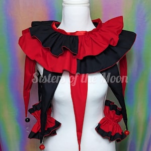 May include: A jester collar featuring alternating red and black fabric panels with ruffled edges. The garment includes long, pointed drapes hanging from the neckline, each finished with a small round bell. The text Sisters of the Moon is visible.