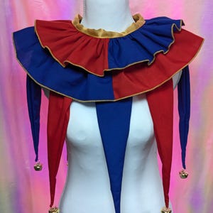 May include: A jester collar featuring a two-toned red and blue fabric design with a gold-colored trim along the edges. The collar includes multiple pointed fabric drapes, each finished with a small metallic bell at the tip, and a gold-colored neck band.