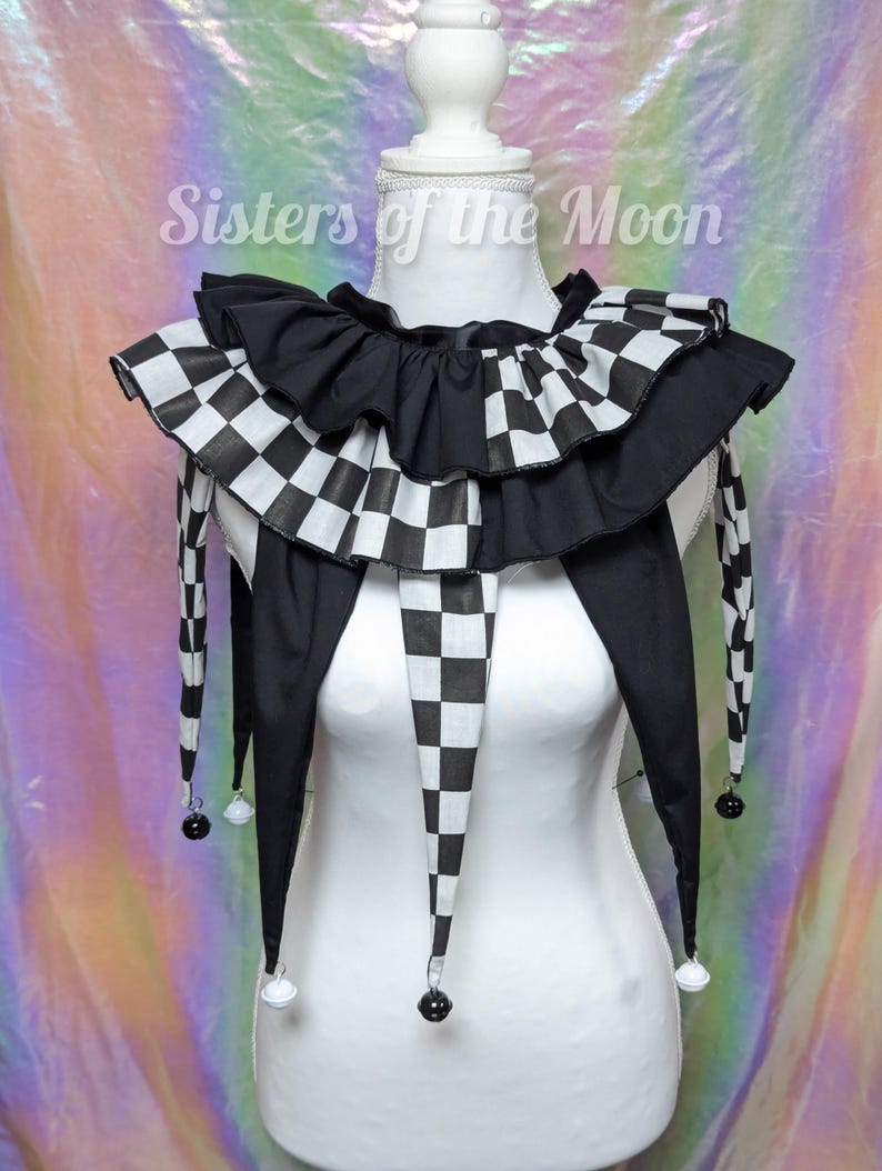 May include: A black and white chequered jester collar with a ruffled design. The collar has long, pointed ends with small bells attached. The text "Sisters of the Moon" is visible in the background.
