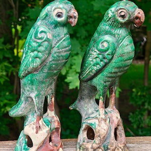 Puede incluir: Dos figuras de loros de cerámica con esmalte verde sobre bases rocosas texturizadas. Cada ave presenta patrones de plumas detallados, un pico curvado y un acabado desgastado con parches de arcilla expuesta bajo el esmalte verde.