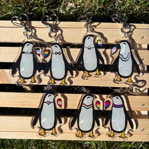 May include: A collection of seven clear acrylic keychains featuring illustrated penguins. Each penguin is decorated with various colorful pride flag patterns on accessories like collars, bow ties, or small heart-shaped charms attached to the keychains.