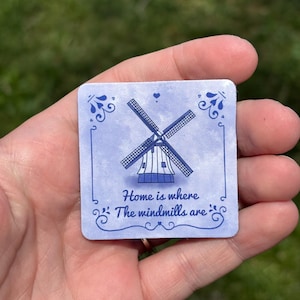 May include: Square blue and white windmill design with the text "Home is where The windmills are". The image features a windmill illustration and decorative floral elements.