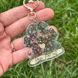 May include: Clear paw-shaped keychain with a gold-colored heart-shaped clasp. The keychain is filled with colorful stars and the words "Dog Parent". A small white heart is in the center of the paw print.