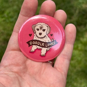 May include: A round, pink button with a cartoon doodle dog illustration. The dog is light brown with a black nose and is wearing a necklace. The button has the words "Doodle Lover" on a banner, with two small hearts on either side.