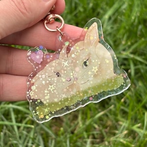 May include: A clear acrylic keychain featuring two light brown bunnies nestled together. The keychain has a gold-colored clasp and is filled with glitter and star-shaped confetti. A pink heart with stars is also visible.