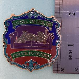 May include: A shield-shaped enamel pin featuring a purple couch with a grey dog lying on it. The text reads "LOYAL ORDER OF" and "COUCH POTATOES" on green banners. A metal ruler next to the pin shows it measures approximately 1.75 inches in height.