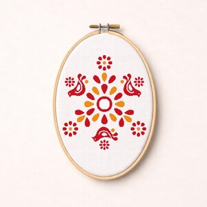 May include: Oval cross-stitch embroidery featuring a symmetrical floral and bird design in red and orange hues. The design is centered on a white fabric, framed by a wooden embroidery hoop. The artwork has a folk art aesthetic.