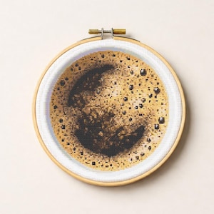 May include: Cross-stitch embroidery of a coffee cup with a detailed depiction of coffee foam and bubbles. The artwork is set within a wooden embroidery hoop, showcasing the texture and color variations of the coffee.