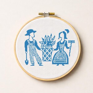以下が含まれることがあります： 青と白のクロスステッチ刺繍枠。植木鉢を持った男性と女性の様式化された描写。男性はシャベルを持ち、女性は熊手を持っています。デザインはフォークアートスタイルです。