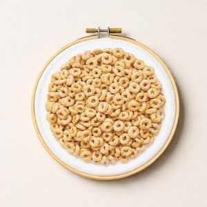 May include: A cross-stitch embroidery hoop featuring a circular design of tan-colored cereal. The cereal pieces are arranged to fill the white fabric within the wooden hoop. The hoop is secured with a brass clasp.