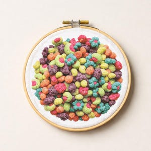 May include: A colorful cross-stitch embroidery hoop featuring a detailed design of various colorful, round shapes. The design includes shades of pink, purple, orange, green, and yellow, resembling a pile of colorful cereal. The embroidery is set in a wooden hoop.