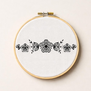 Puede incluir: Bordado de punto de cruz terminado en un bastidor de madera. El diseño presenta un patrón floral simétrico en hilo negro sobre tela blanca. Las flores estilizadas y las hojas crean un borde decorativo. El bastidor es de madera clara.