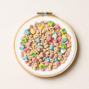 May include: Cross-stitch embroidery of a bowl of cereal with colorful marshmallow shapes. The embroidery is set in a wooden embroidery hoop. The cereal pieces are tan, with pink, blue, yellow, and green marshmallows.
