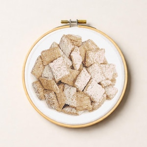Può includere: Un telaio da ricamo a punto croce con un mucchio di cereali quadrati color marrone chiaro. I cereali sono su uno sfondo bianco e il telaio è in legno con una chiusura in metallo.