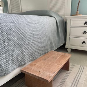 May include: A rectangular wooden step stool with a natural light brown finish and visible wood grain. The stool features a flat top surface constructed from two joined planks with visible circular indentations and two sturdy side supports.