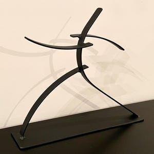 May include: A black metal sculpture depicting a stylized figure in motion. The sculpture has a rectangular base and curved lines suggesting arms and legs. The figure appears to be running or dancing, with outstretched arms.