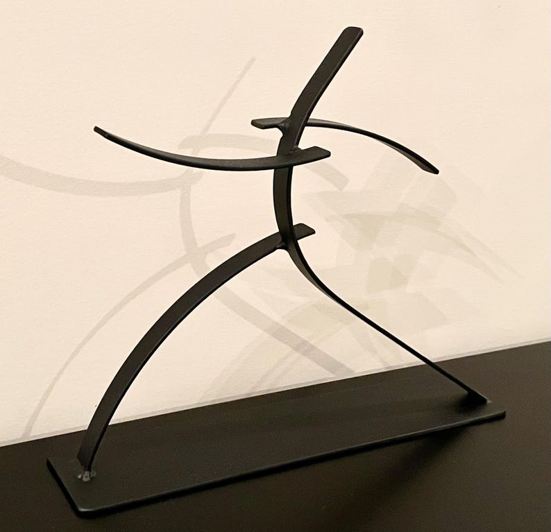 May include: A black metal sculpture depicting a stylized figure in motion. The sculpture has a rectangular base and curved lines suggesting arms and legs. The figure appears to be running or dancing, with outstretched arms.