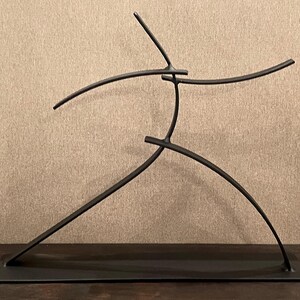 May include: A black metal sculpture depicting a stylized human figure in motion. The sculpture has a minimalist design with curved and straight lines, mounted on a rectangular base. The figure appears to be mid-stride or dancing.